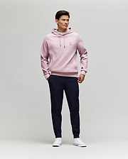 A man stands against a plain light background, wearing Psycho Bunny's MENS BEN TECH JOGGER PULL ON - MB0600013 in dark navy, paired with a light pink hoodie and white sneakers. He smiles slightly, hands in pockets, looking to the side. (4)