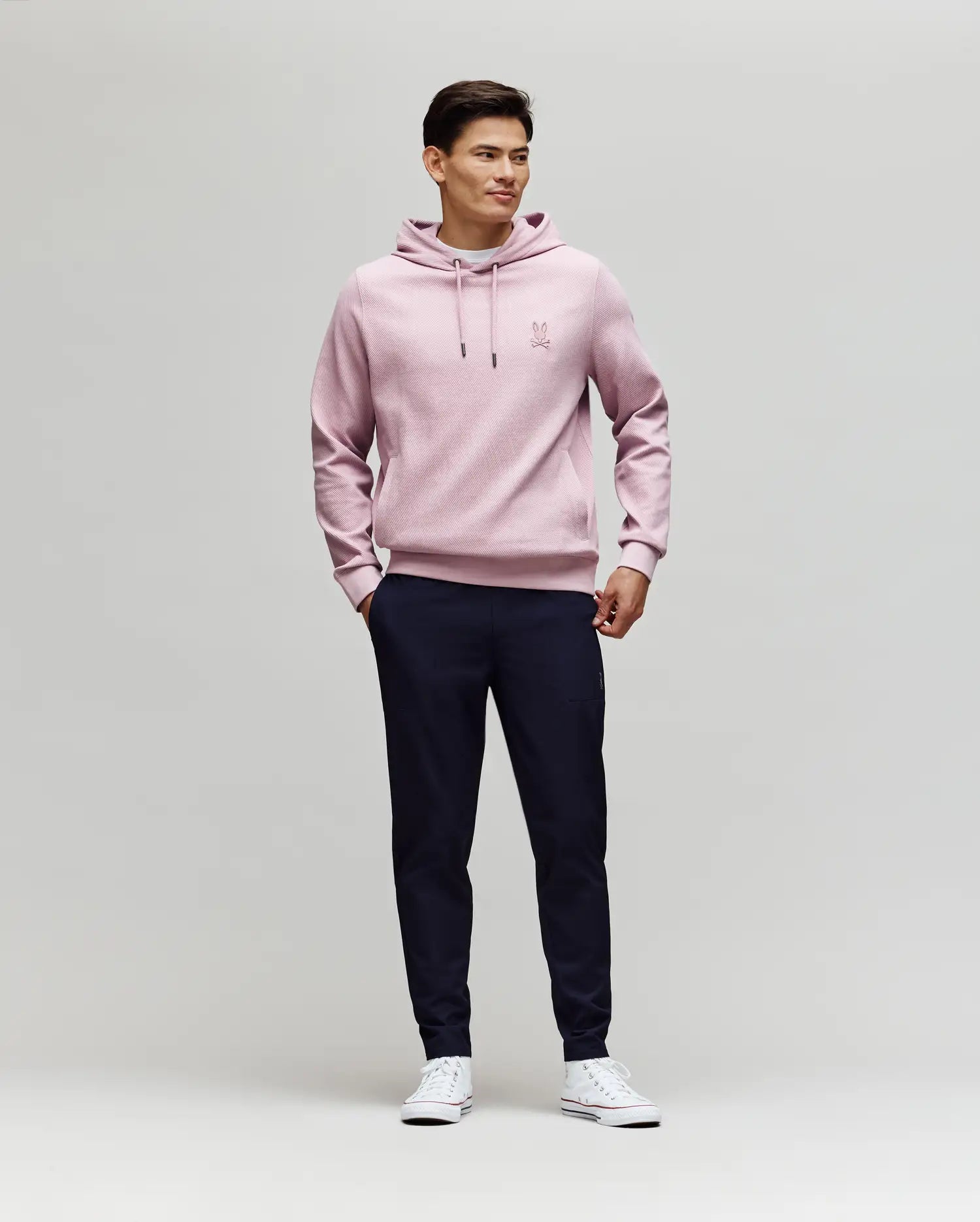 A man stands against a plain light background, wearing Psycho Bunny's MENS BEN TECH JOGGER PULL ON - MB0600013 in dark navy, paired with a light pink hoodie and white sneakers. He smiles slightly, hands in pockets, looking to the side. (4)