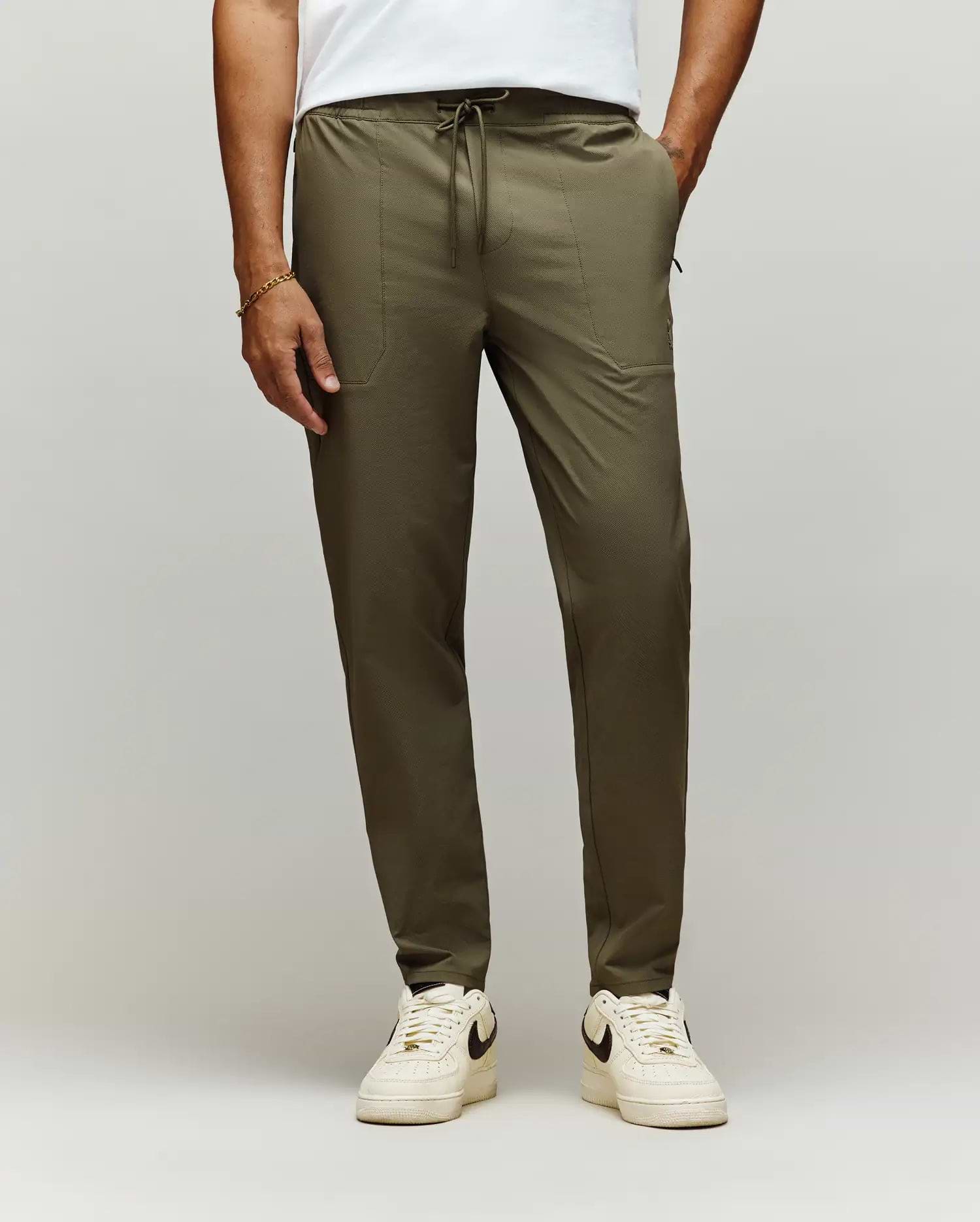 A man stands against a plain light background, showing only his lower body in Psycho Bunny MENS BEN TECH JOGGER PULL ON - MB0600013 (olive green), paired with a white t-shirt and white Nike sneakers. (1)