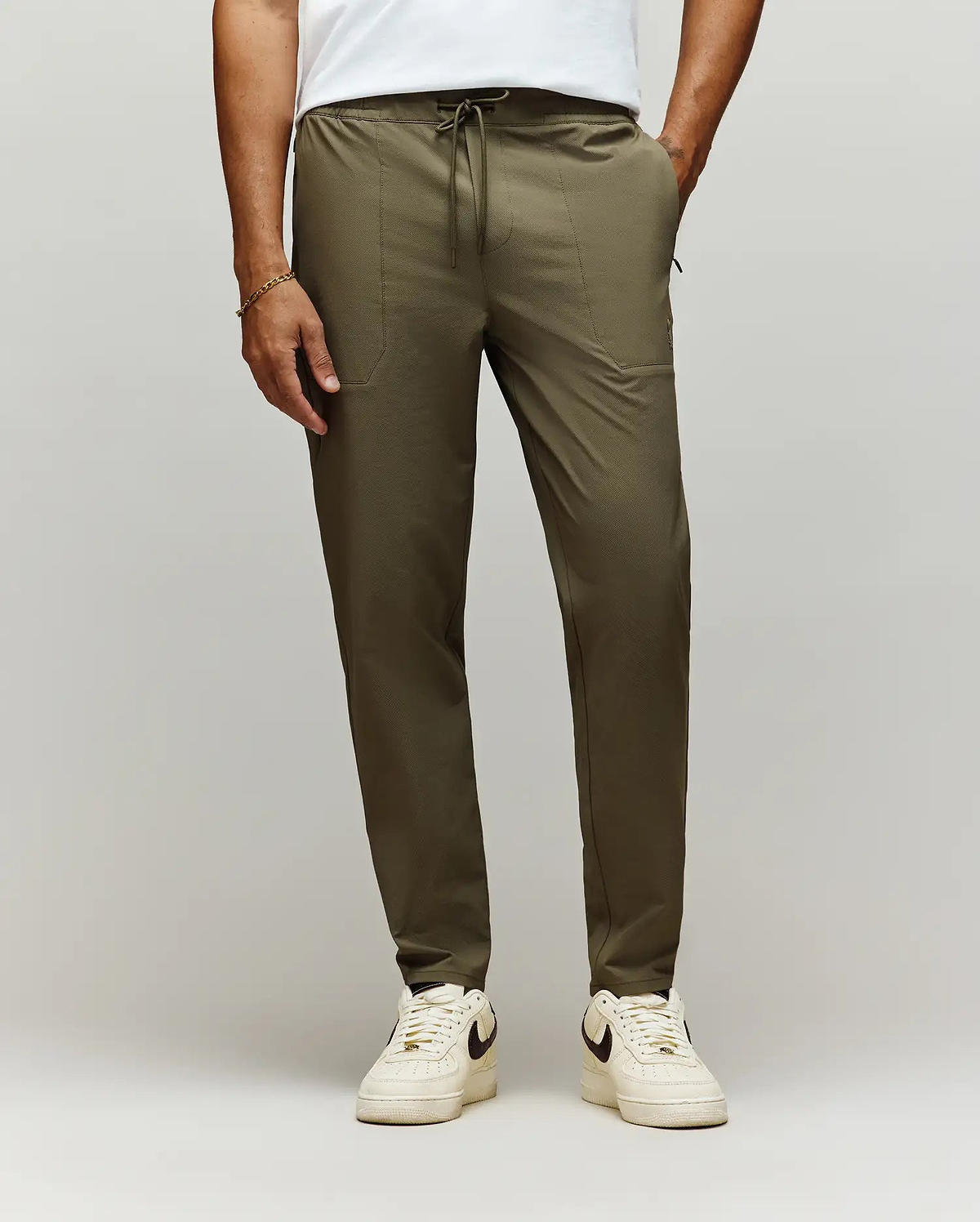 A man stands against a plain light background, showing only his lower body in Psycho Bunny MENS BEN TECH JOGGER PULL ON - MB0600013 (olive green), paired with a white t-shirt and white Nike sneakers. (1)