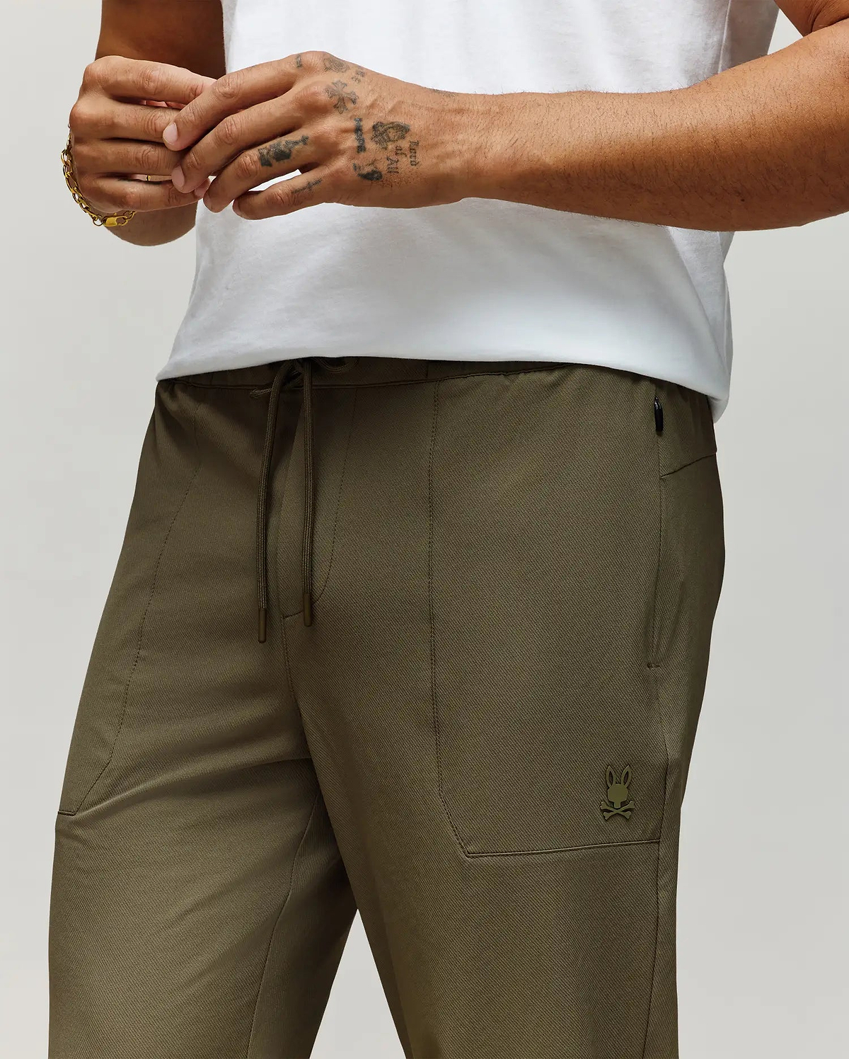 A person wears Psycho Bunny's MENS BEN TECH JOGGER PULL ON (MB0600013) in olive green, featuring a polyester-spandex blend, drawstring waist, and small bunny logo, paired with a white shirt; only torso/upper legs and one tattooed hand with bracelet are visible. (2)