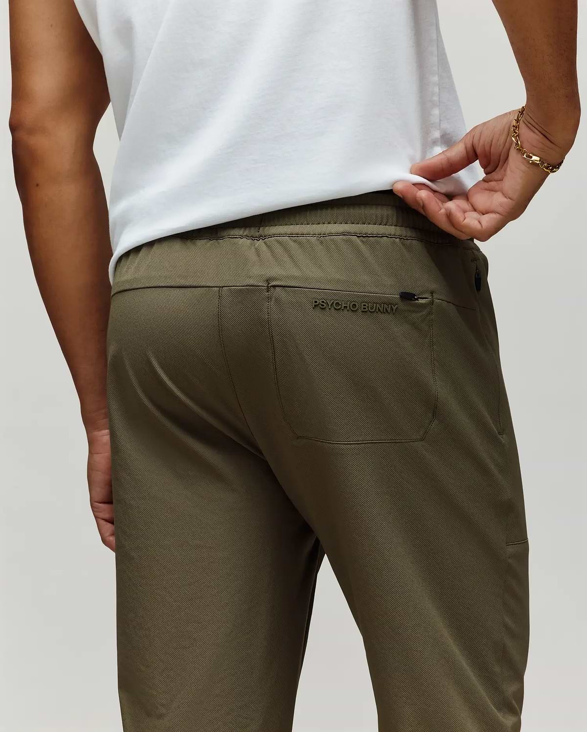 A man is shown from the back wearing Psycho Bunny's MENS BEN TECH JOGGER PULL ON (MB0600013) in olive green, featuring a rear pocket with "Psycho Bunny" embroidery, paired with a white shirt and a gold bracelet on his left wrist. (3)