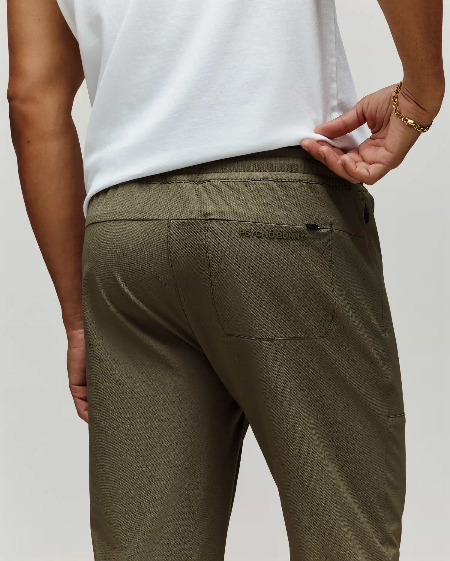 A man is shown from the back wearing Psycho Bunny's MENS BEN TECH JOGGER PULL ON (MB0600013) in olive green, featuring a rear pocket with "Psycho Bunny" embroidery, paired with a white shirt and a gold bracelet on his left wrist. (3)