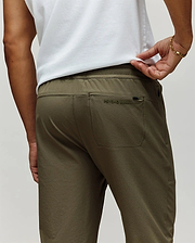 A man is shown from the back wearing Psycho Bunny's MENS BEN TECH JOGGER PULL ON (MB0600013) in olive green, featuring a rear pocket with "Psycho Bunny" embroidery, paired with a white shirt and a gold bracelet on his left wrist. (3)