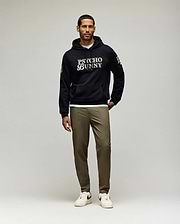 A man with short dark hair and a trimmed beard, looking slightly left, wears a black Psycho Bunny hoodie and olive green Psycho Bunny MENS BEN TECH JOGGER PULL ON - MB0600013 pants, paired with white sneakers. (4)