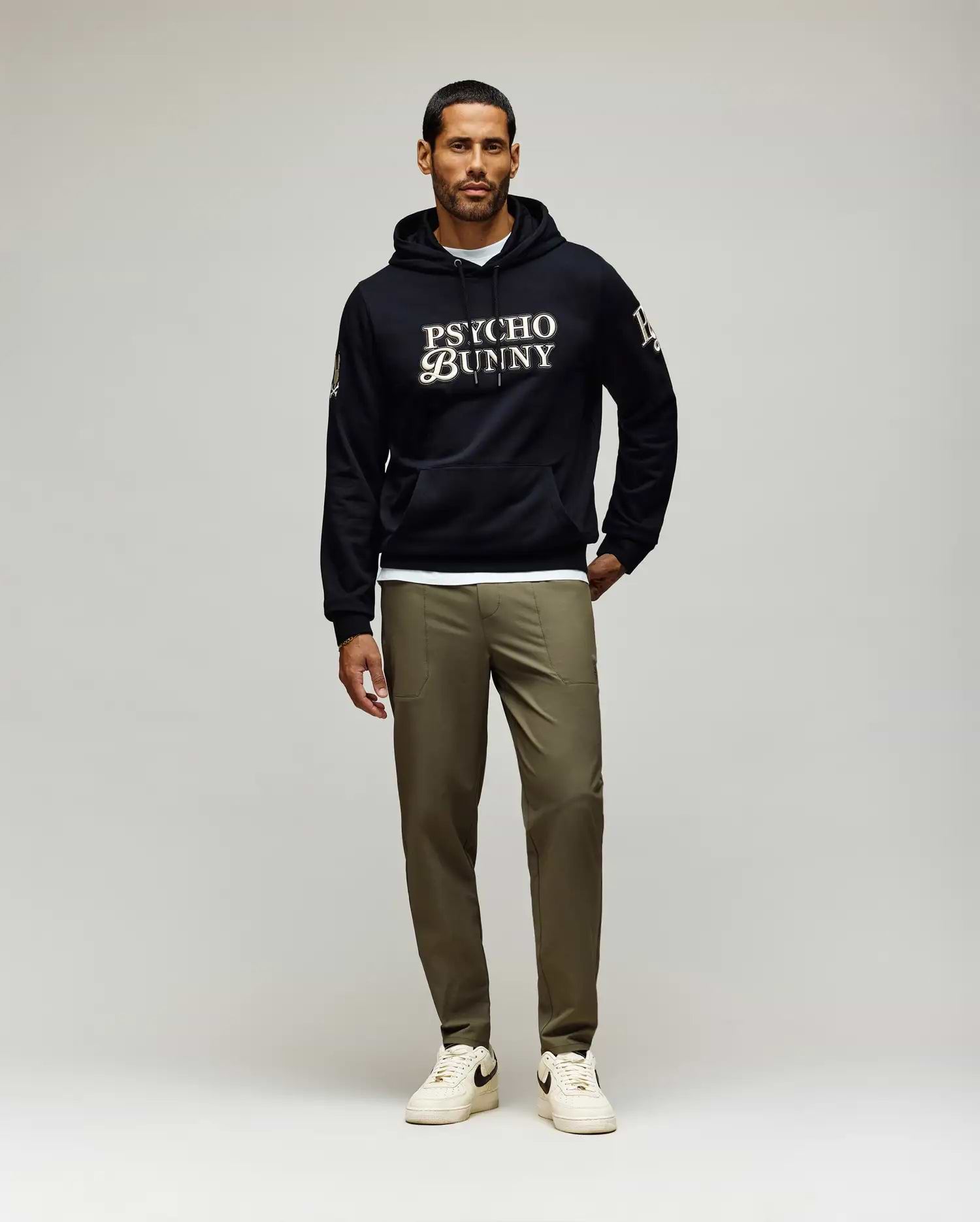 A man with short dark hair and a trimmed beard, looking slightly left, wears a black Psycho Bunny hoodie and olive green Psycho Bunny MENS BEN TECH JOGGER PULL ON - MB0600013 pants, paired with white sneakers. (4)
