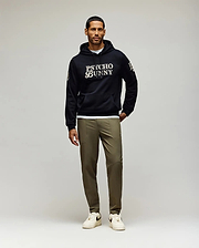 A man with short dark hair and a trimmed beard, looking slightly left, wears a black Psycho Bunny hoodie and olive green Psycho Bunny MENS BEN TECH JOGGER PULL ON - MB0600013 pants, paired with white sneakers. (4)