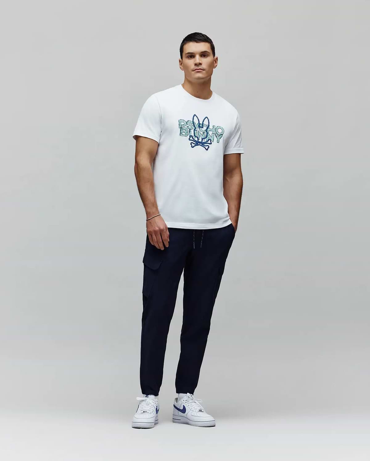 White jersey t-shirt with a large blue gradient Psycho Bunny skull graphic on the chest, paired with navy Mens Luka Cargo Pant featuring side pockets and a tapered fit. (5)