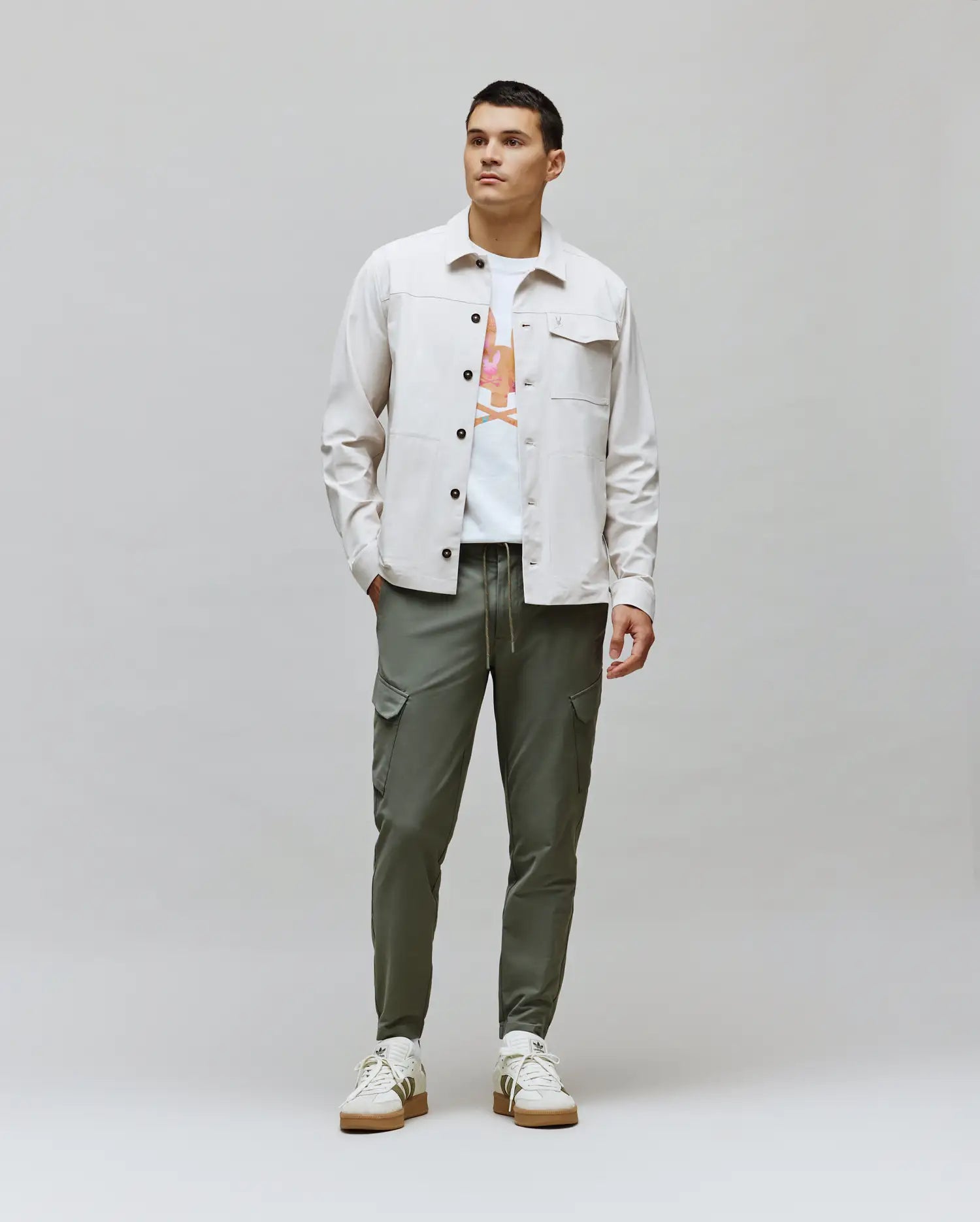 Light beige twill shirt jacket with chest pockets layered over a white classic fit jersey tee featuring a vibrant Psycho Bunny skull graphic, paired with slim fit olive green Mens Luka Cargo Pant. (5)