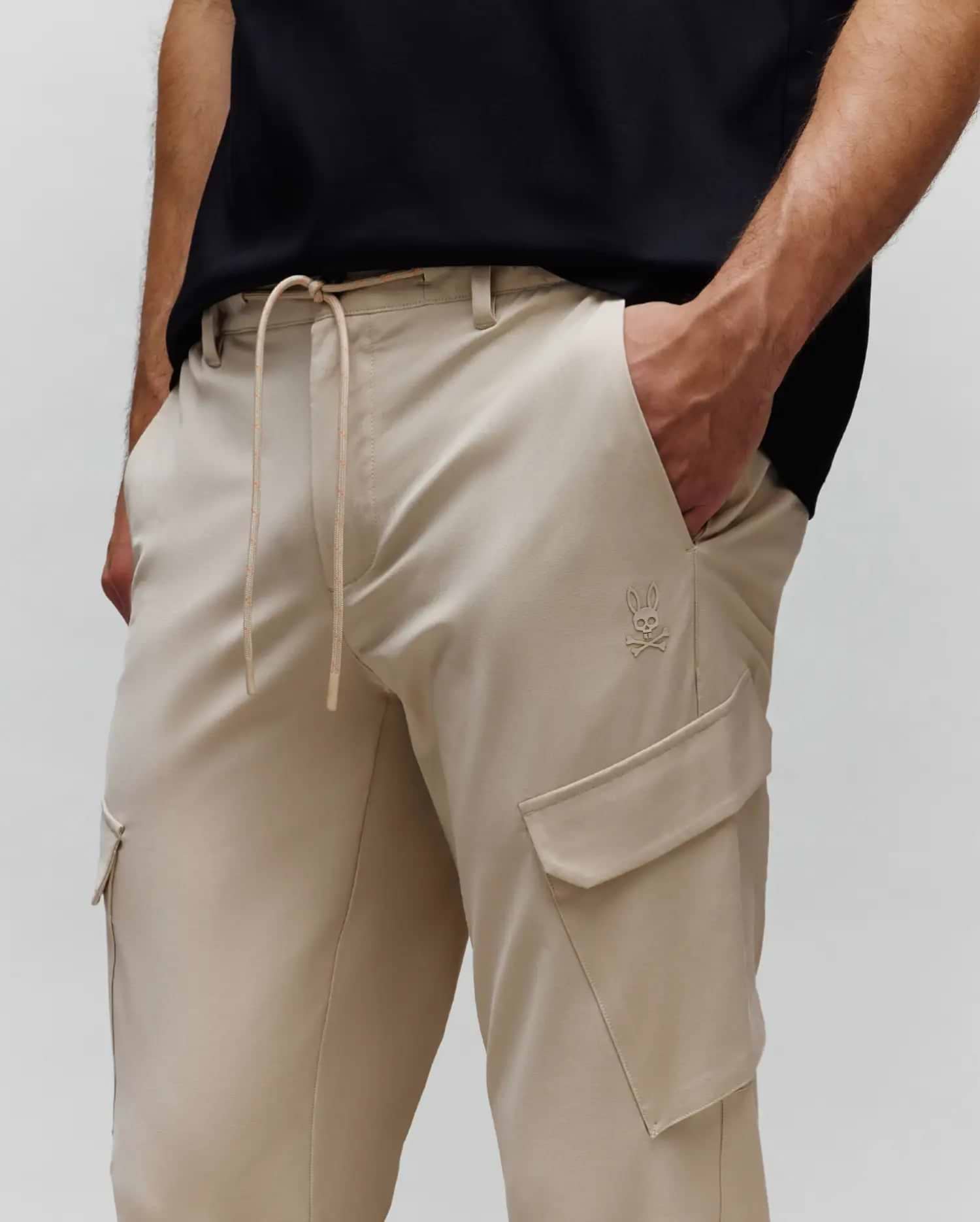 Mens Luka Cargo Pant in khaki, featuring a relaxed fit, smooth fabric, drawstring waist with subtle orange flecks, and a small embroidered Psycho Bunny skull logo on the left cargo pocket.