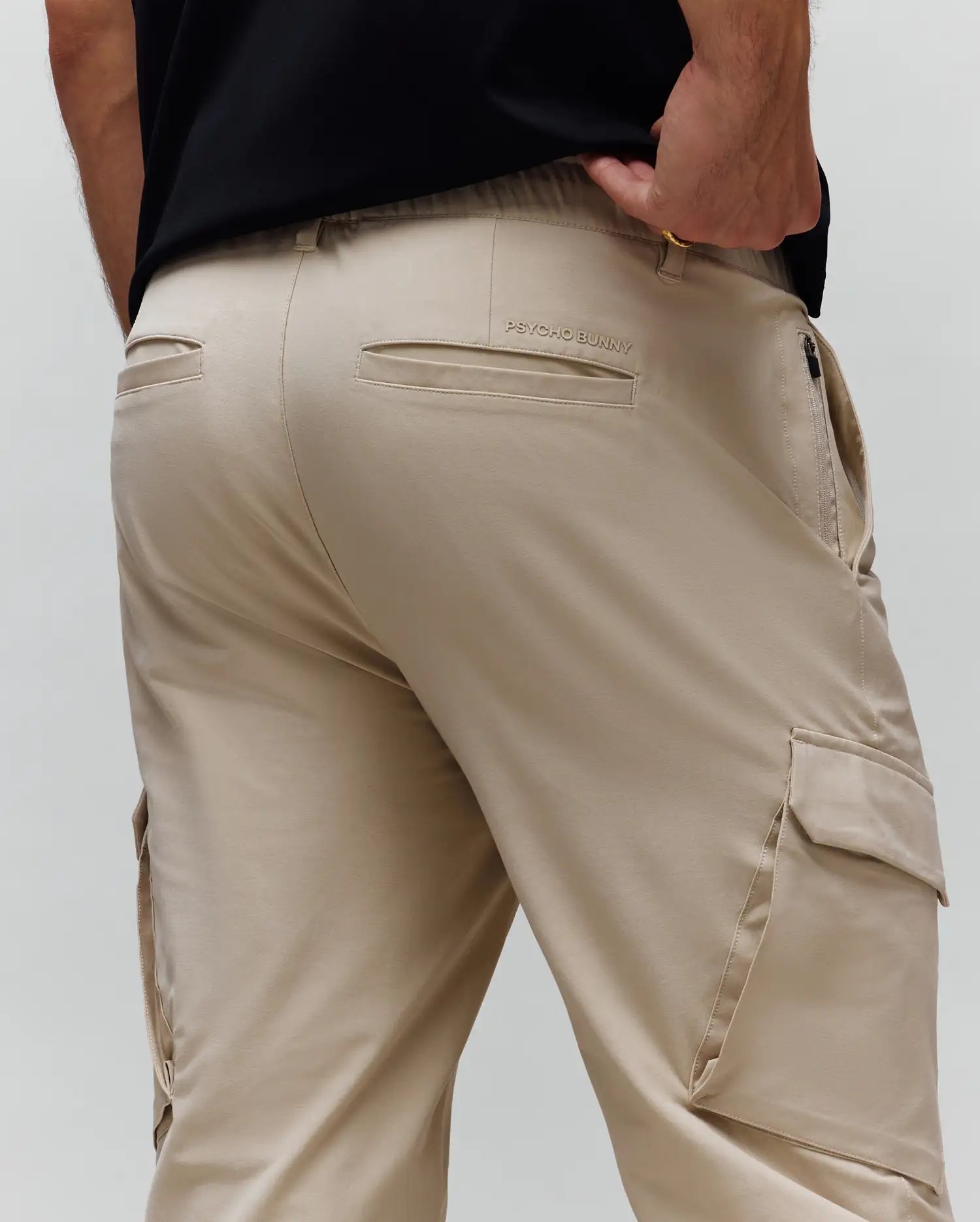 Mens Luka Cargo Pant in light tan, featuring a relaxed fit, smooth fabric, side cargo pockets, back welt pockets, elastic waistband, and a subtle embroidered Psycho Bunny text logo.