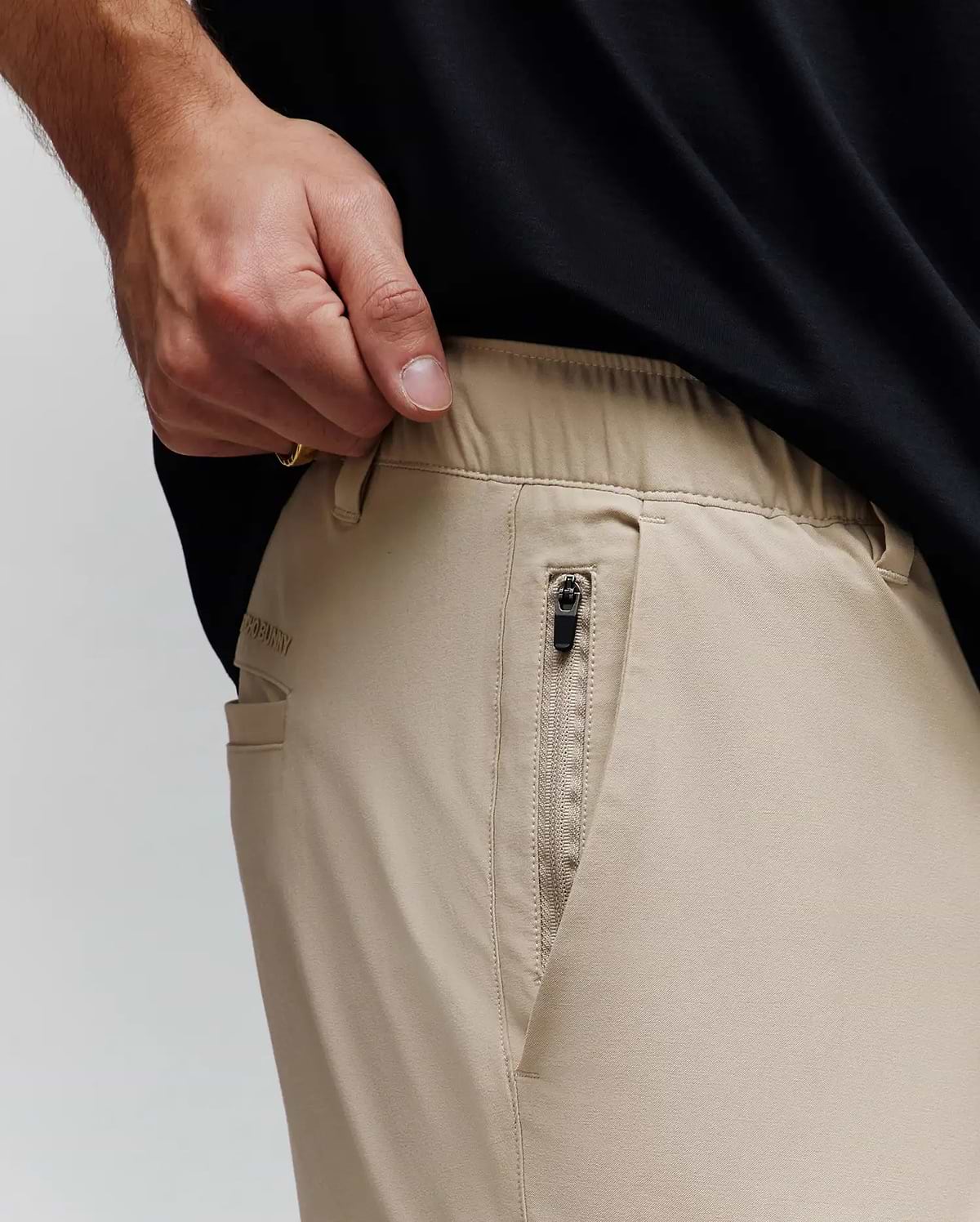 Mens Luka Cargo Pant in khaki, a relaxed fit with smooth fabric, elastic waistband, zippered side pocket with black pull, and subtle Psycho Bunny text logo on the side.