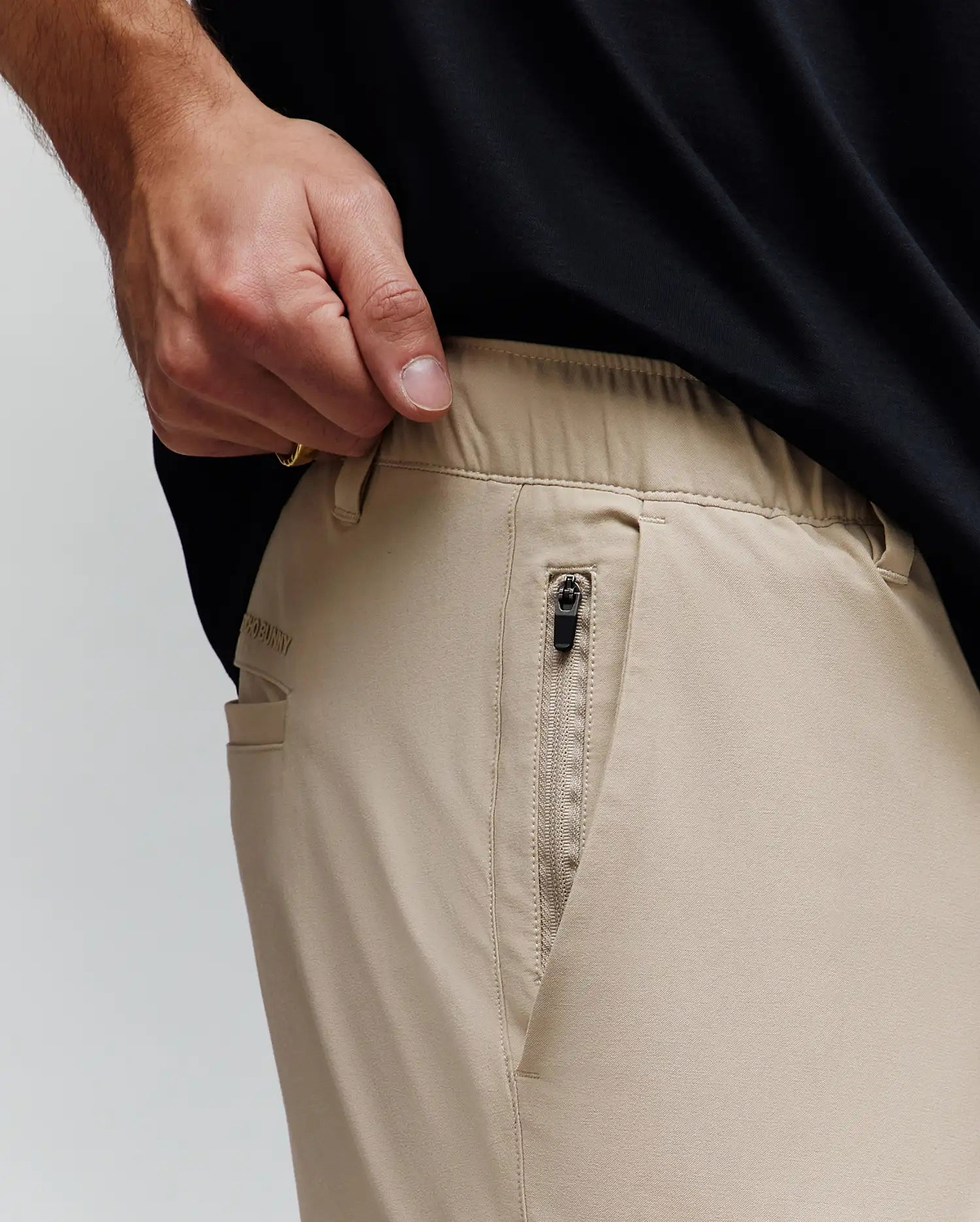 Mens Luka Cargo Pant in khaki, a relaxed fit with smooth fabric, elastic waistband, zippered side pocket with black pull, and subtle Psycho Bunny text logo on the side.