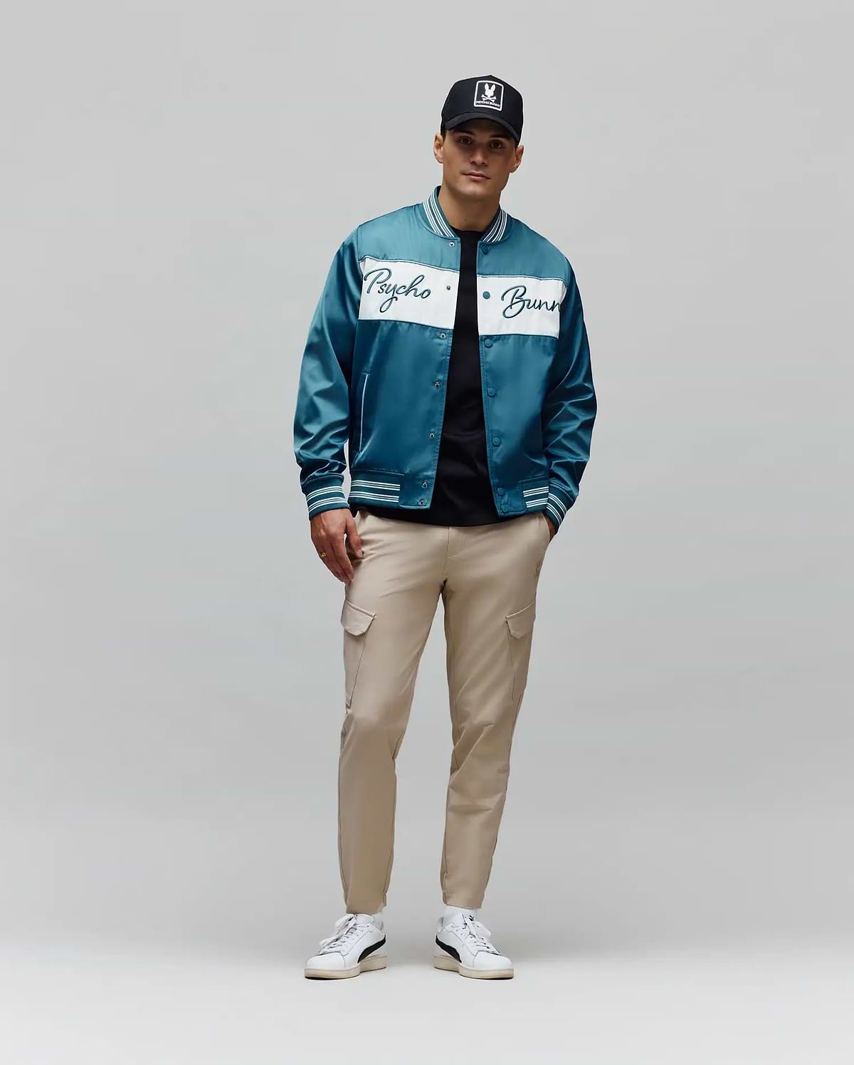 Teal satin bomber jacket with white Psycho Bunny script across chest and striped cuffs, over a black tee, paired with slim-fit Khaki Mens Luka Cargo Pant and a black Psycho Bunny skull logo cap.