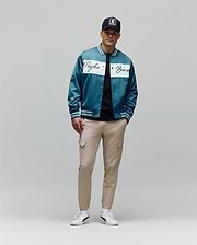 Teal satin bomber jacket with white Psycho Bunny script across chest and striped cuffs, over a black tee, paired with slim-fit Khaki Mens Luka Cargo Pant and a black Psycho Bunny skull logo cap.