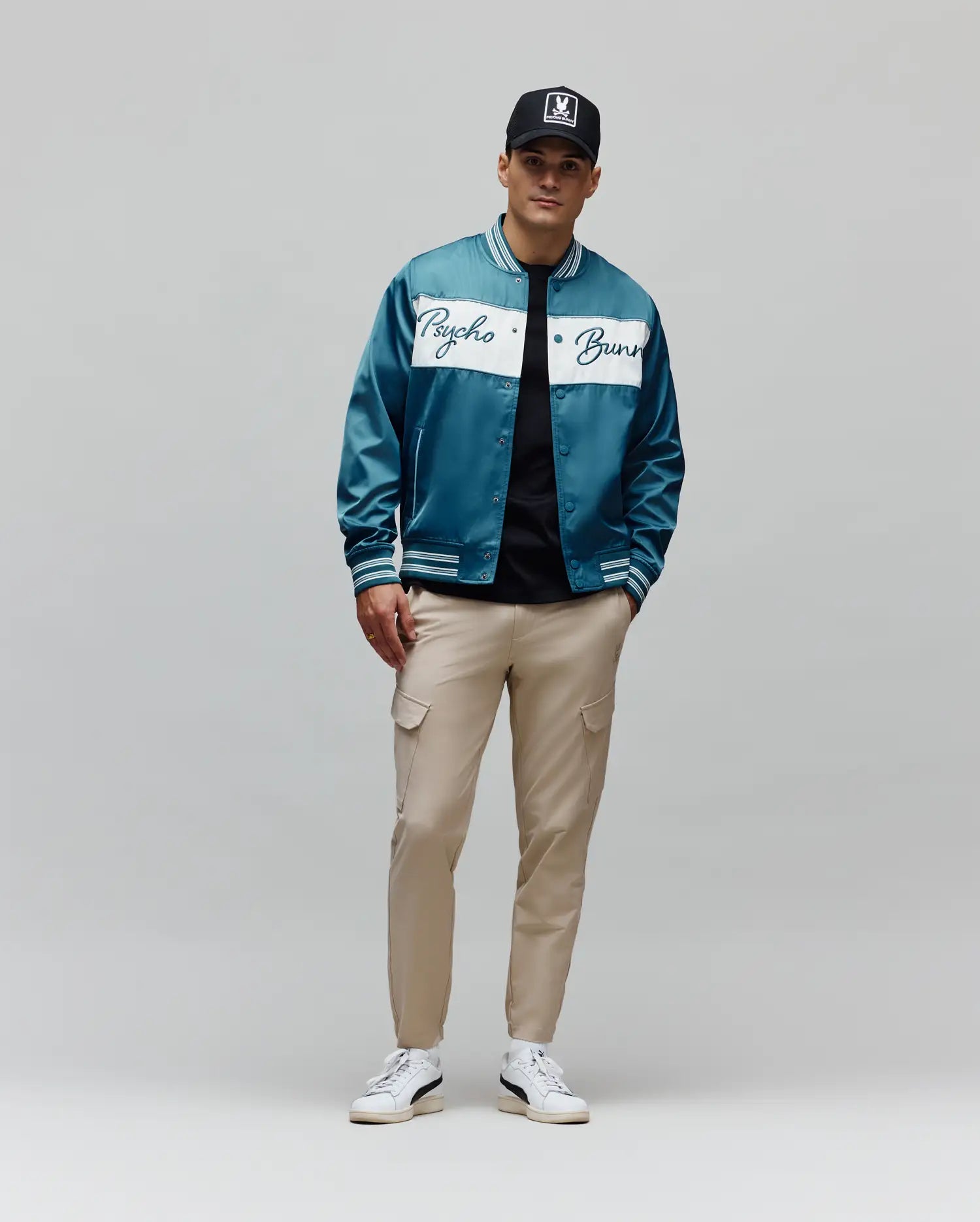 Teal satin bomber jacket with white Psycho Bunny script across chest and striped cuffs, over a black tee, paired with slim-fit Khaki Mens Luka Cargo Pant and a black Psycho Bunny skull logo cap.