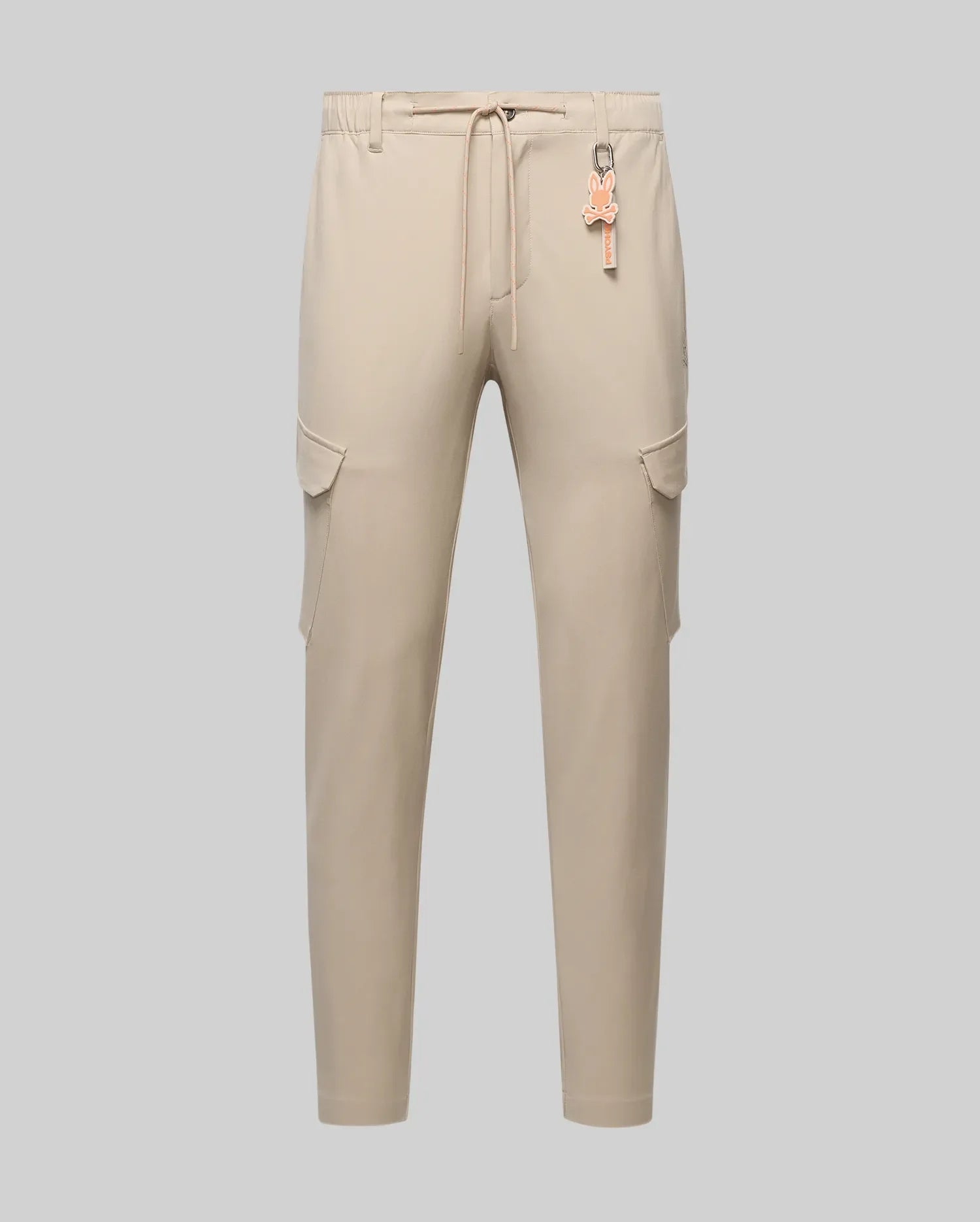 Mens Luka Cargo Pant in beige, featuring a relaxed fit, smooth fabric, side cargo pockets, and an elastic drawstring waist with an attached orange Psycho Bunny skull logo keychain.