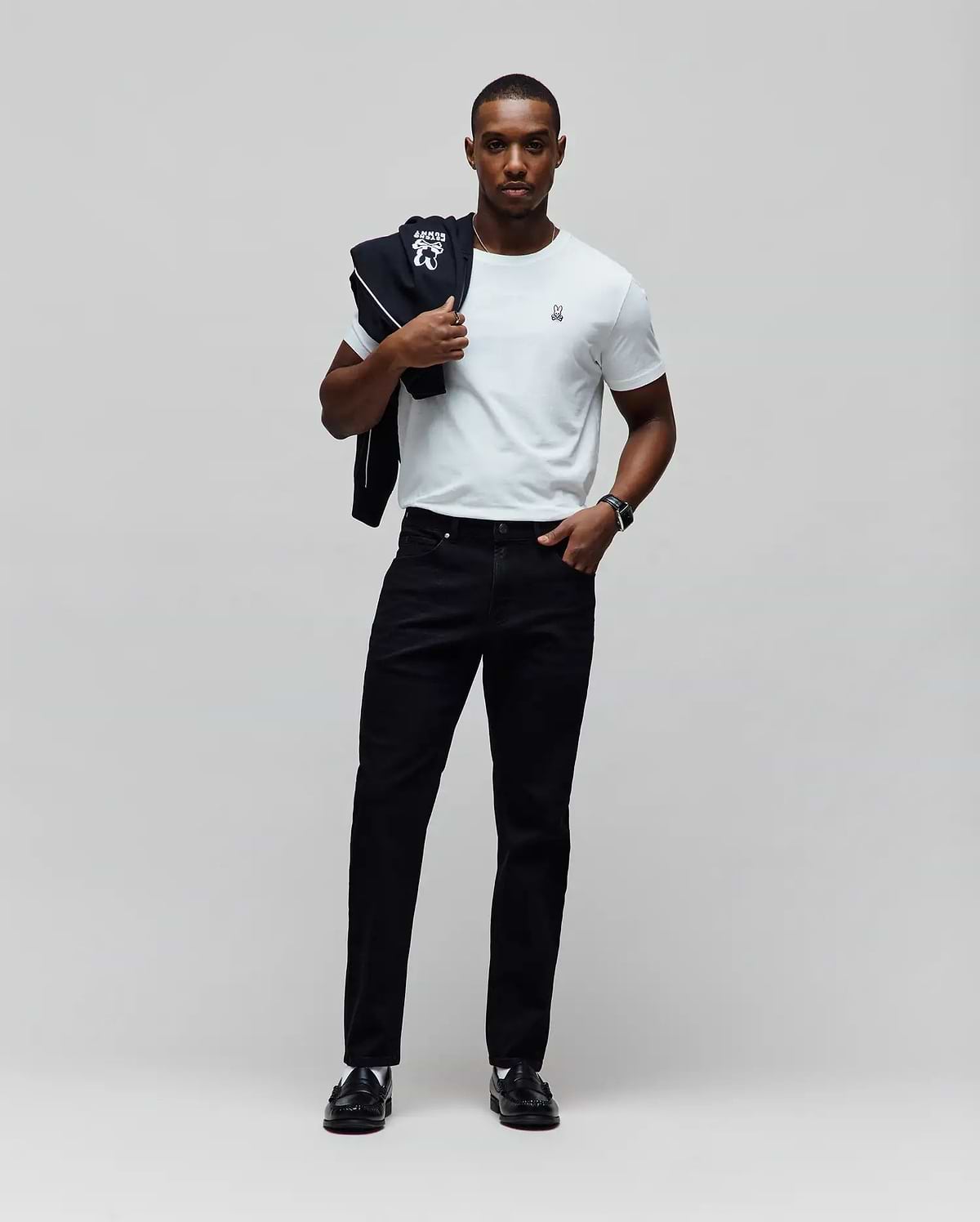 White jersey t-shirt with small embroidered Psycho Bunny logo on left chest, paired with black Mens Emerson Slim Fit Denim Pant and a black track jacket with large white Psycho Bunny graphic over the shoulder. (5)