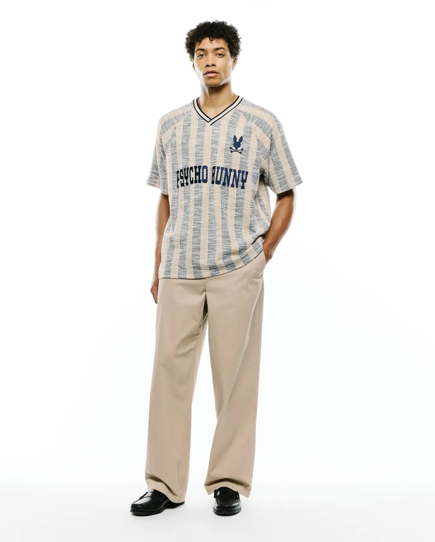 Textured beige and blue vertical striped knit V-neck shirt with black trim, featuring large PSYCHO BUNNY text and small skull logo on chest, paired with relaxed fit khaki Mens Aidan Chino Pant.