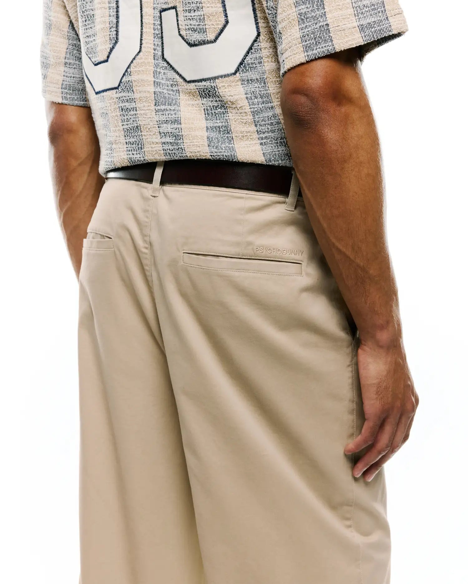 MENS Aidan Chino Pant in classic beige twill, featuring a relaxed fit and subtle PSYCHOBUNNY embroidery above the back pocket, paired with a textured beige and navy striped shirt with a large white graphic.