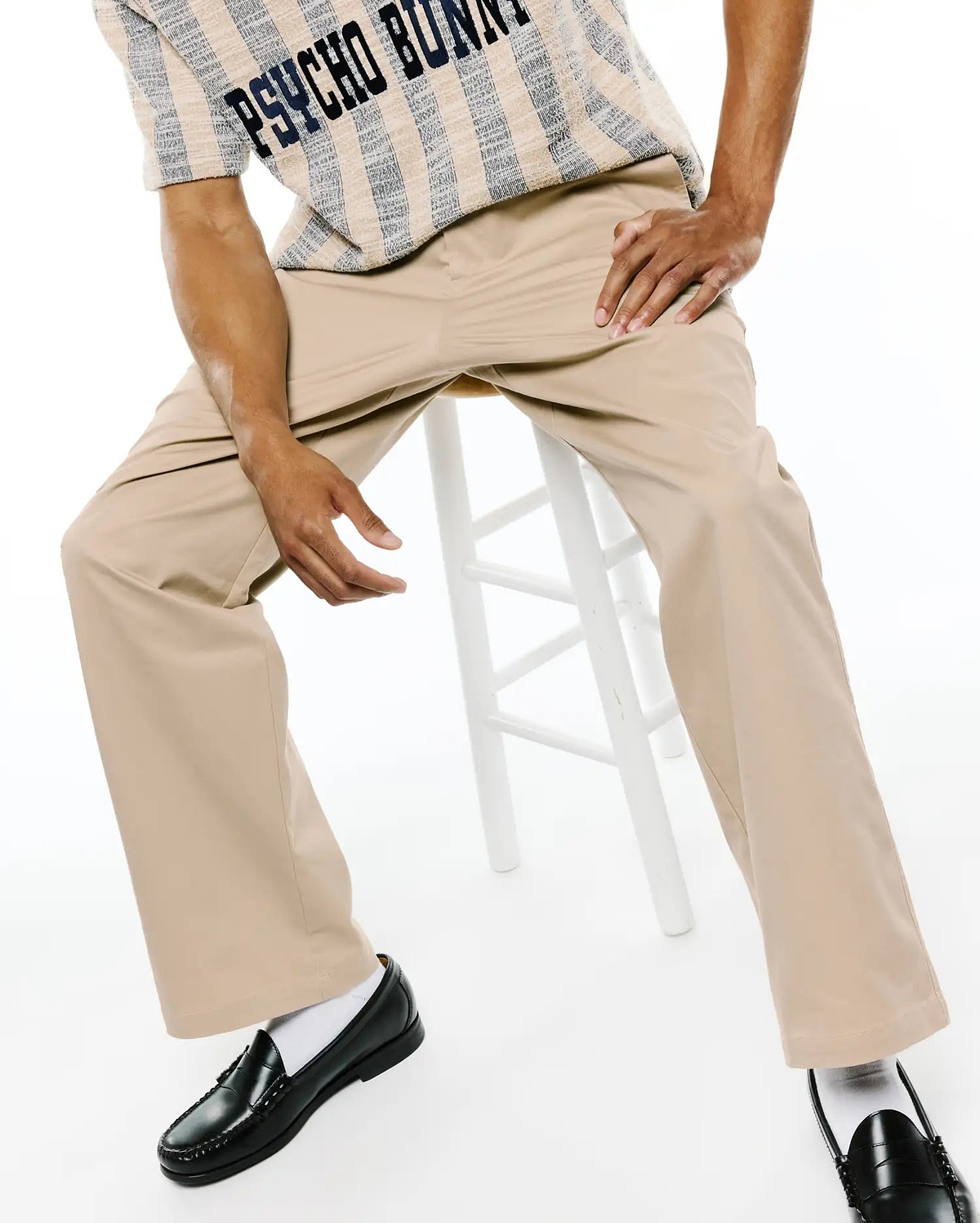 MENS AIDAN CHINO PANT in relaxed fit khaki twill, paired with a cream and navy striped textured knit short-sleeve shirt featuring large PSYCHO BUNNY text across the chest.