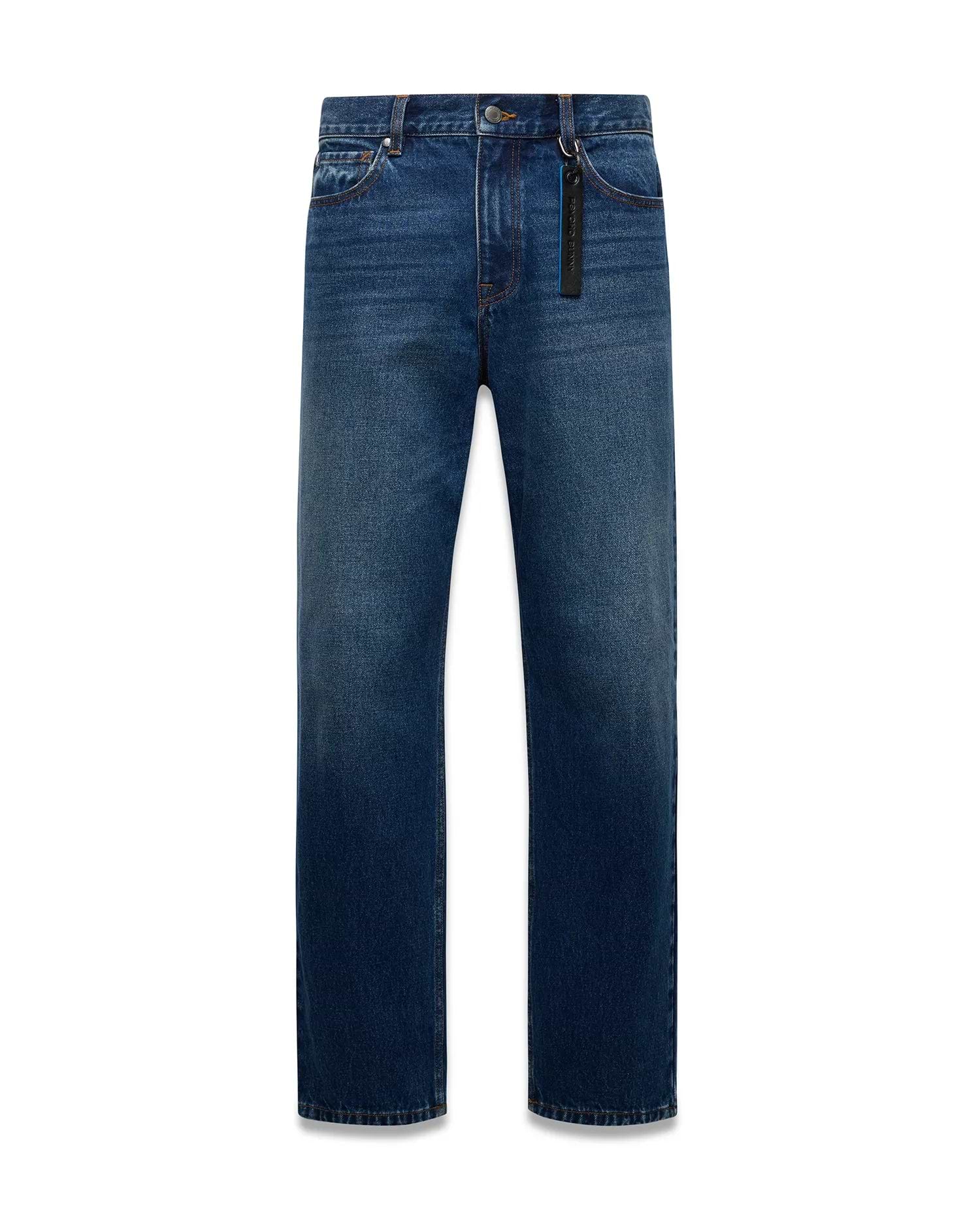 Mens Beck Denim Relaxed Pant in medium blue denim with a faded wash, orange contrast stitching, and a black Psycho Bunny branded tag with blue text on the front belt loop.