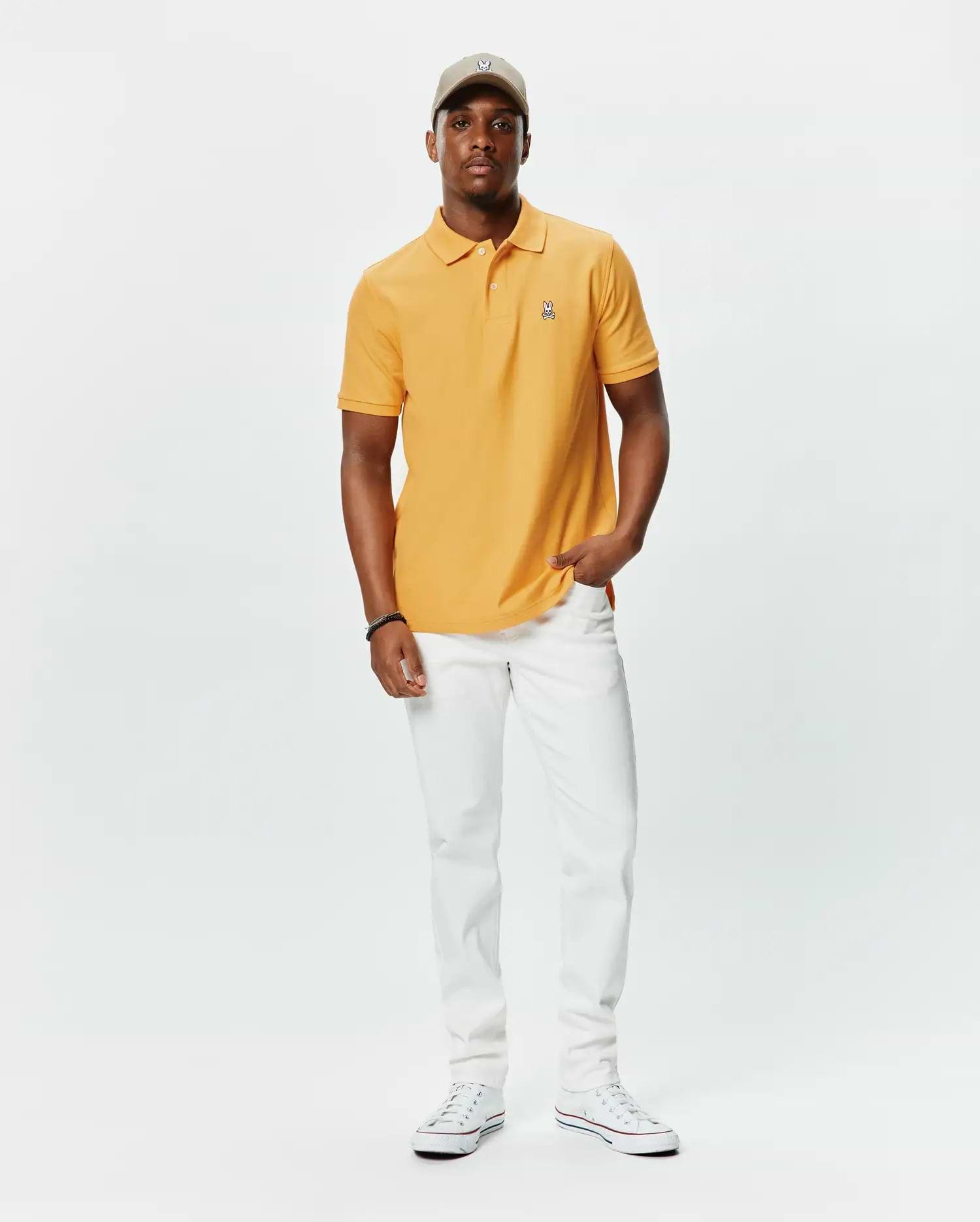 Orange pique polo shirt with classic fit and embroidered Psycho Bunny skull logo on left chest, paired with white slim-fit Mens Emerson Denim Pant and a beige cap with matching logo.