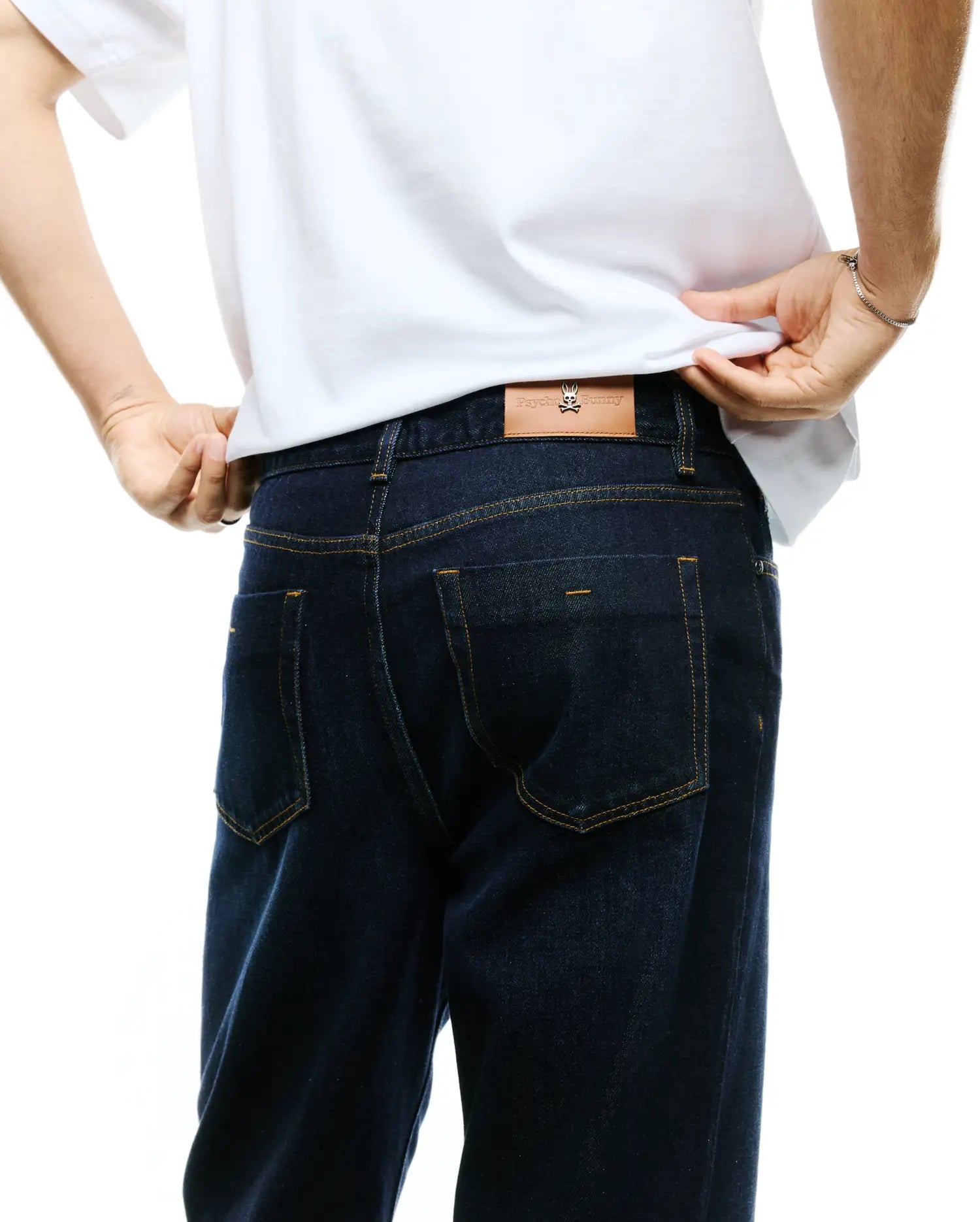 Men's Beck Oversized Denim Pants in dark indigo denim, featuring tan contrast stitching, classic back pockets, and a brown leather Psycho Bunny skull logo patch on the rear waistband. (2)