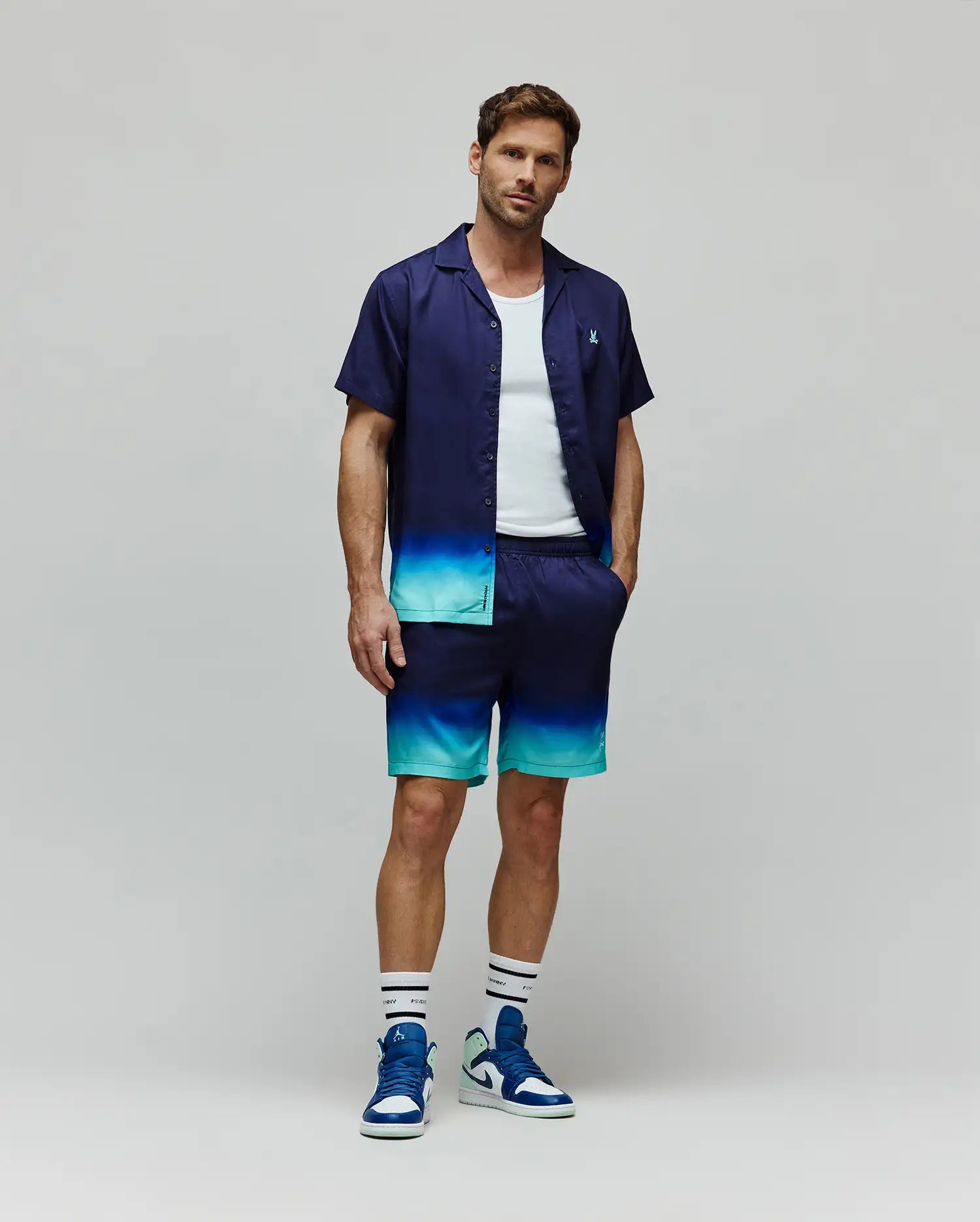 Relaxed fit navy short-sleeve button-up shirt and Mens Dax Tencel Short, with a vibrant blue and teal ombre fade. Shirt has a small embroidered Psycho Bunny skull logo, shorts include Psycho Bunny branding.