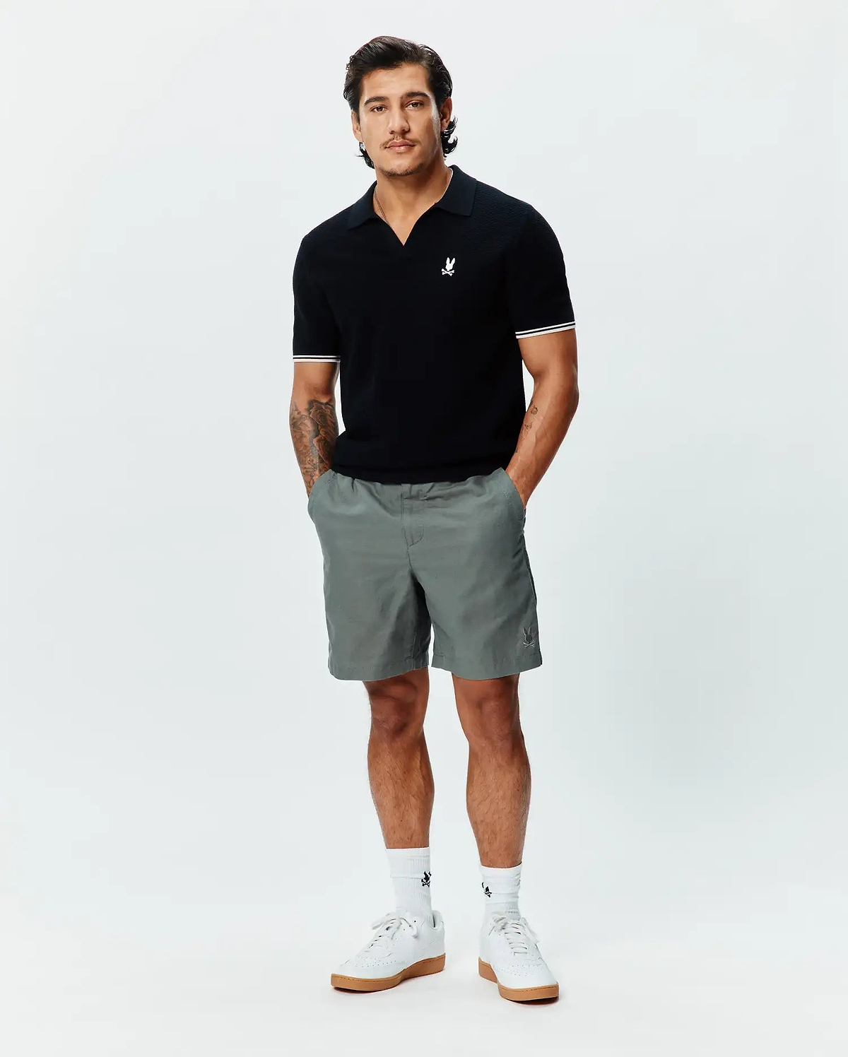 Black knit polo with an open collar, white striped cuffs, and a small white embroidered Psycho Bunny logo. Paired with grey Mens Ivan Linen Short, featuring a tonal embroidered bunny logo.