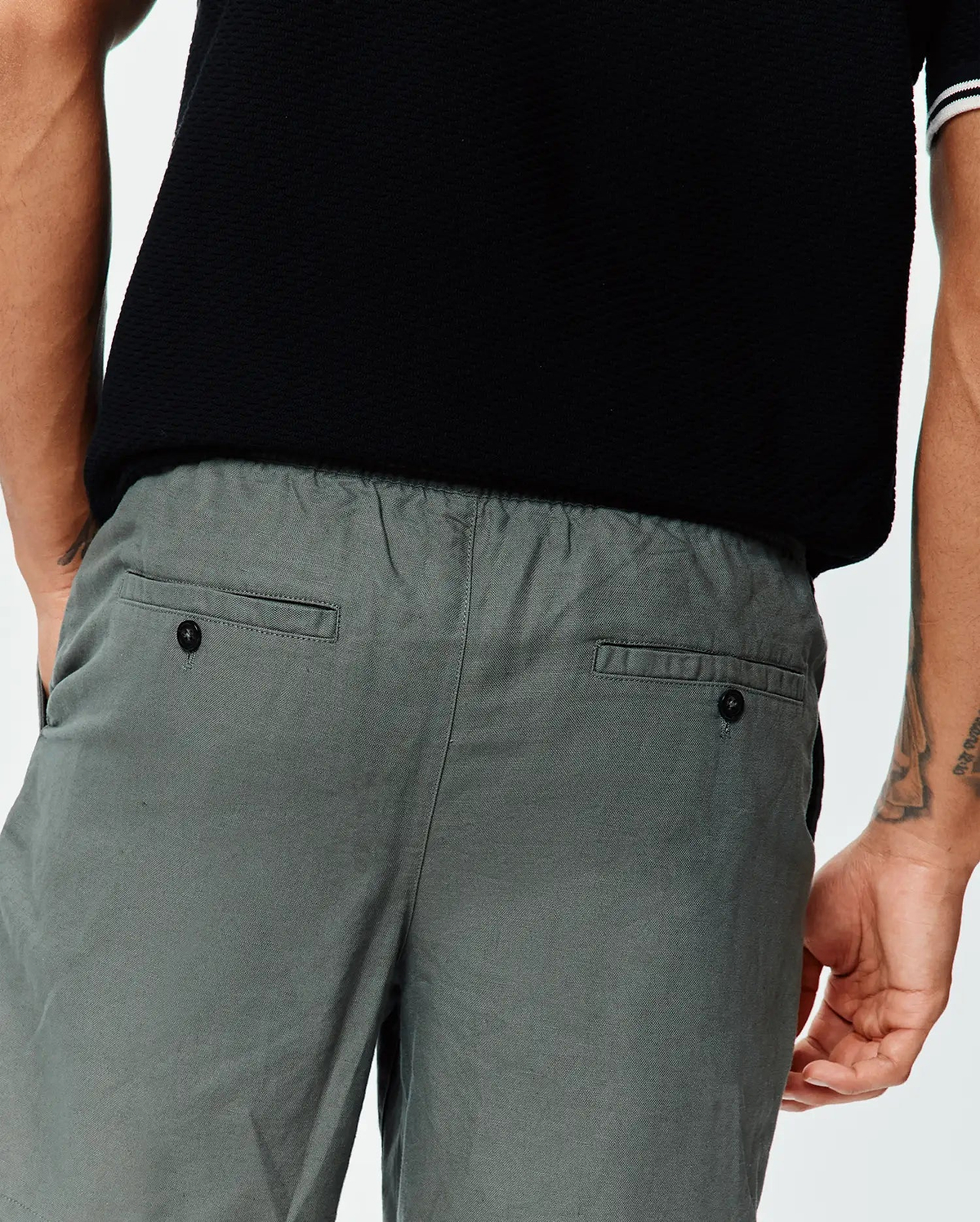 Mens Ivan Linen Short in relaxed grey with an elastic waist and buttoned back welt pockets, paired with a classic black textured pique polo featuring white and black striped cuffs.