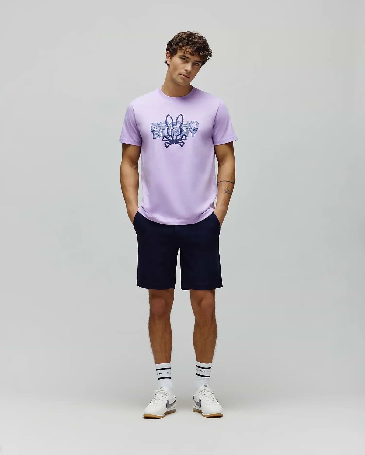 Lavender jersey t-shirt with a large distressed blue Psycho Bunny skull logo and text graphic on the chest, paired with classic fit navy MENS SHIRO TWILL SHORT.