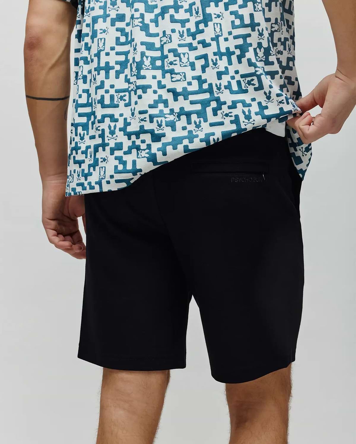 Black mens shiro twill short, classic fit, with subtle embroidered Psycho Bunny text on the back right pocket, complemented by a white and teal pixelated Psycho Bunny logo print shirt. (3)