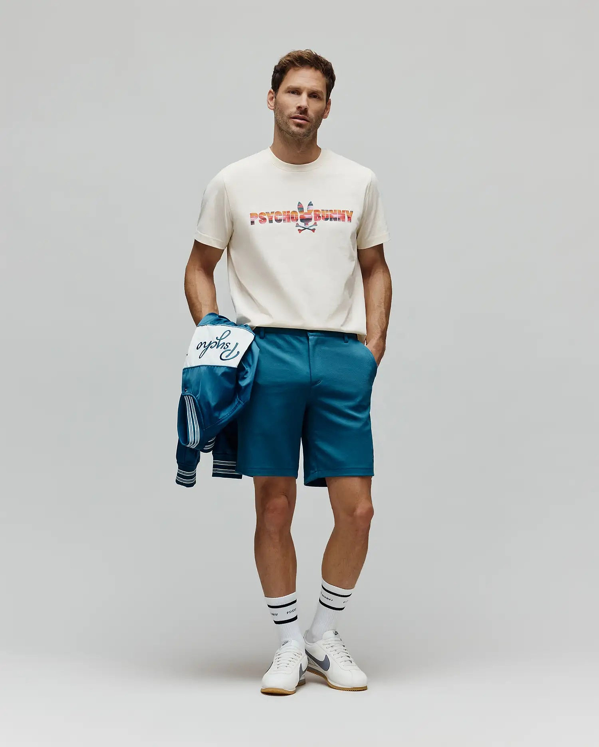 Classic fit cream jersey t-shirt featuring a large, colorful patterned Psycho Bunny logo, paired with Mens Shiro Twill Short in teal, and a matching teal bomber jacket with white Psycho script. (4)