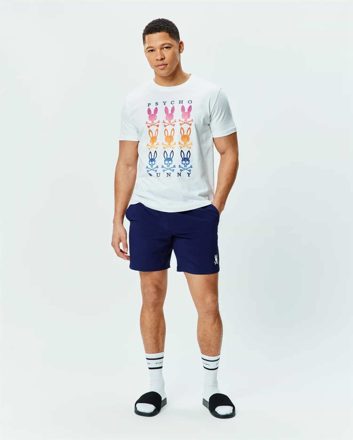 White jersey t-shirt with a large central graphic of nine gradient Psycho Bunny skull logos and PSYCHO BUNNY text, paired with navy Mens Blaze Sport Short with a small white embroidered Psycho Bunny logo on the left leg.