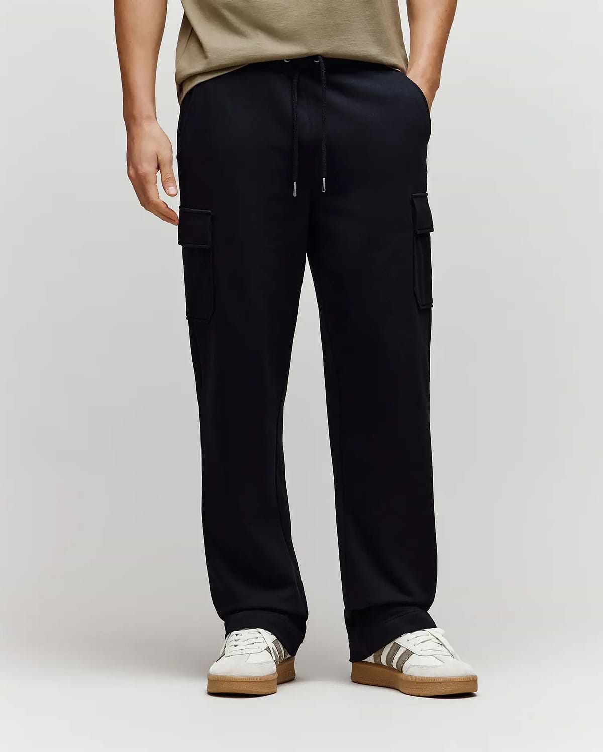 Wearing Psycho Bunny MENS NASH CARGO SWEATPANT - MB0800010, black with roomy cargo pockets and sweatpant comfort, paired with white/beige sneakers and a beige shirt. The background is light; only the lower body is fully visible.