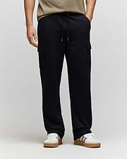 Wearing Psycho Bunny MENS NASH CARGO SWEATPANT - MB0800010, black with roomy cargo pockets and sweatpant comfort, paired with white/beige sneakers and a beige shirt. The background is light; only the lower body is fully visible.