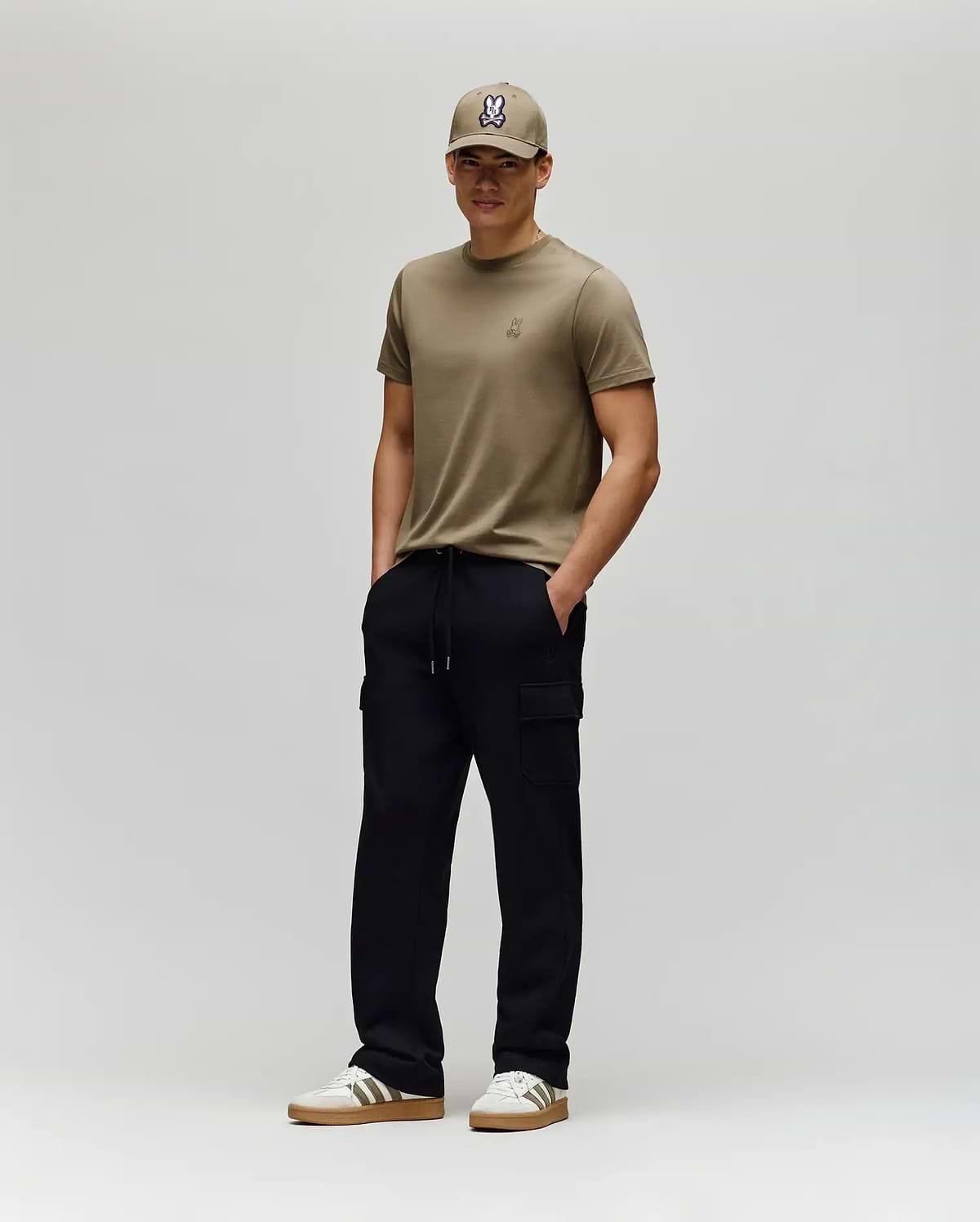 A man stands against a plain light gray background, wearing a khaki T-shirt, matching cap, white sneakers with brown soles, and Psycho Bunny MENS NASH CARGO SWEATPANT - MB0800010. His hands are in his pockets and he smiles slightly.