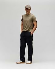 A man stands against a plain light gray background, wearing a khaki T-shirt, matching cap, white sneakers with brown soles, and Psycho Bunny MENS NASH CARGO SWEATPANT - MB0800010. His hands are in his pockets and he smiles slightly.