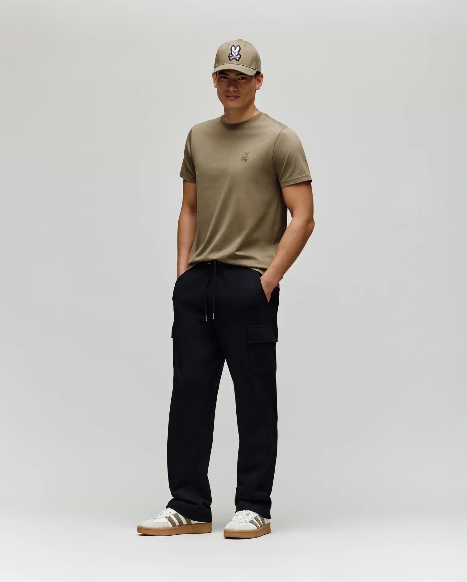 A man stands against a plain light gray background, wearing a khaki T-shirt, matching cap, white sneakers with brown soles, and Psycho Bunny MENS NASH CARGO SWEATPANT - MB0800010. His hands are in his pockets and he smiles slightly.