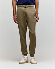 A person wears Psycho Bunny MENS WADE CORD PATCH SWEATPANT - MB0800014 in olive green, featuring "PSY BNK" on the left thigh, rib knit cuffs, and an athletic fit, paired with a navy shirt and white sneakers against a plain background.