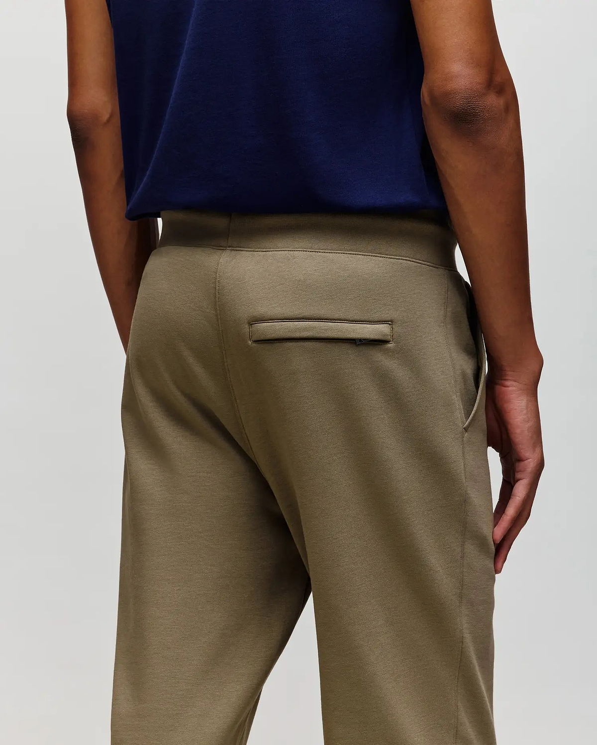 A person wears Psycho Bunny MENS WADE CORD PATCH SWEATPANT - MB0800014 in olive green, made from a premium cotton blend, shown from the waist down (back view) with one hand at their side and a single welt back pocket visible.