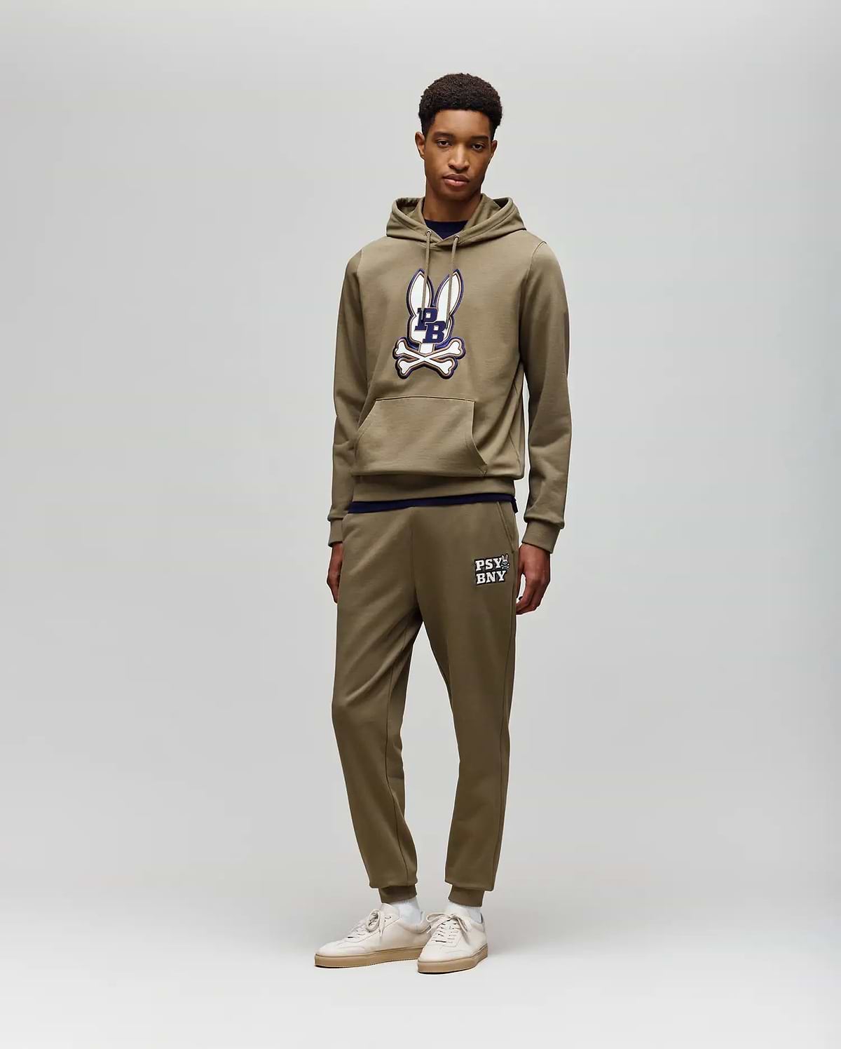 A young man stands against a light gray background, wearing Psycho Bunny's MENS WADE CORD PATCH SWEATPANT (MB0800014) with "PSYCHO BUNNY" text, rib knit cuffs, and an olive hoodie for an athletic style.