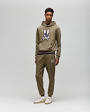 A young man stands against a light gray background, wearing Psycho Bunny's MENS WADE CORD PATCH SWEATPANT (MB0800014) with "PSYCHO BUNNY" text, rib knit cuffs, and an olive hoodie for an athletic style.