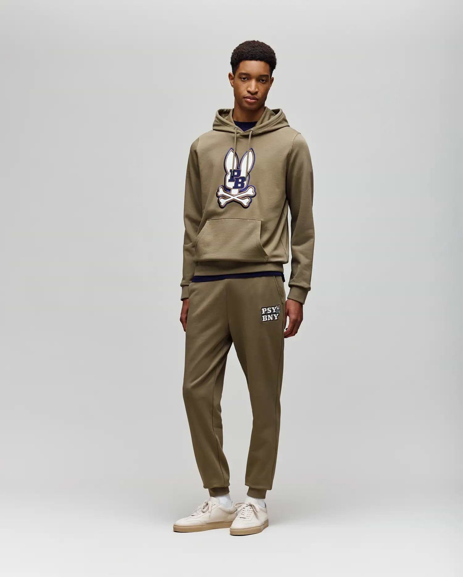 A young man stands against a light gray background, wearing Psycho Bunny's MENS WADE CORD PATCH SWEATPANT (MB0800014) with "PSYCHO BUNNY" text, rib knit cuffs, and an olive hoodie for an athletic style.