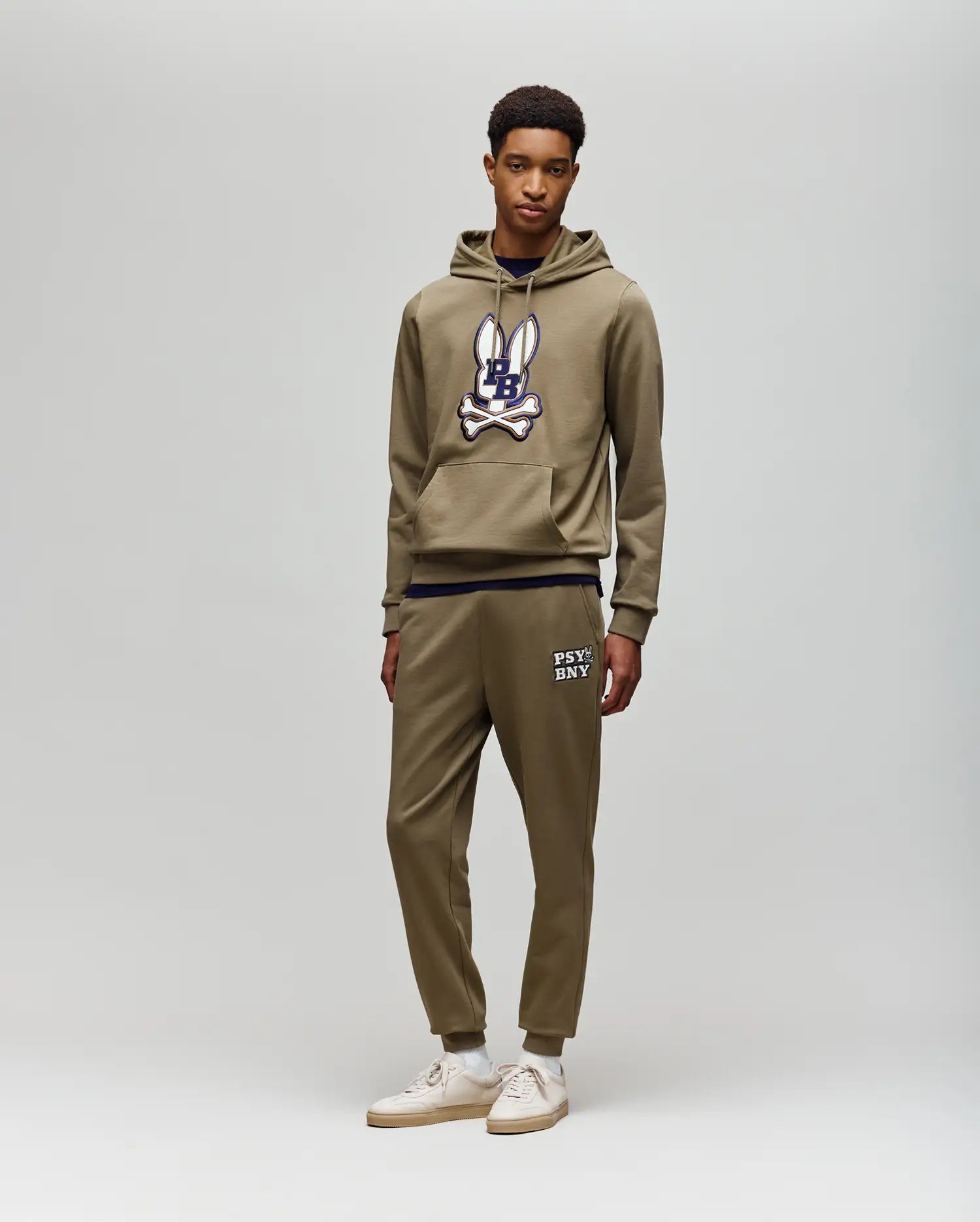 A young man stands against a light gray background, wearing Psycho Bunny's MENS WADE CORD PATCH SWEATPANT (MB0800014) with "PSYCHO BUNNY" text, rib knit cuffs, and an olive hoodie for an athletic style.