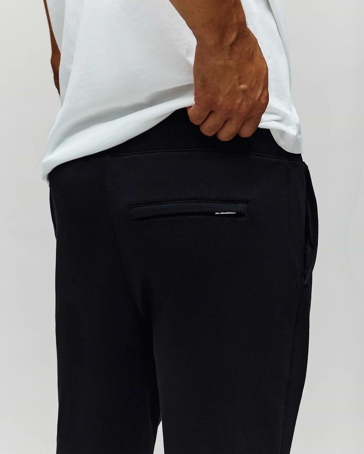 A person wearing a white T-shirt and Psycho Bunny MENS AUGUSTE JOGGER - MB0800015 is shown from behind, hand near the waistband. The back pocket and the 100% cotton fabric texture are visible.