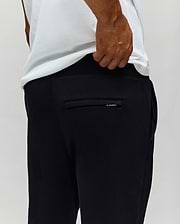 A person wearing a white T-shirt and Psycho Bunny MENS AUGUSTE JOGGER - MB0800015 is shown from behind, hand near the waistband. The back pocket and the 100% cotton fabric texture are visible.