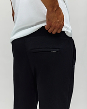 A person wearing a white T-shirt and Psycho Bunny MENS AUGUSTE JOGGER - MB0800015 is shown from behind, hand near the waistband. The back pocket and the 100% cotton fabric texture are visible.