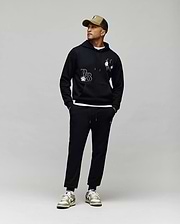 A man in a tan cap, black hoodie with white chenille patches, Psycho Bunny MENS AUGUSTE JOGGER - MB0800015 (100% cotton), and white sneakers with gold accents stands against a light gray background, looking to his right.
